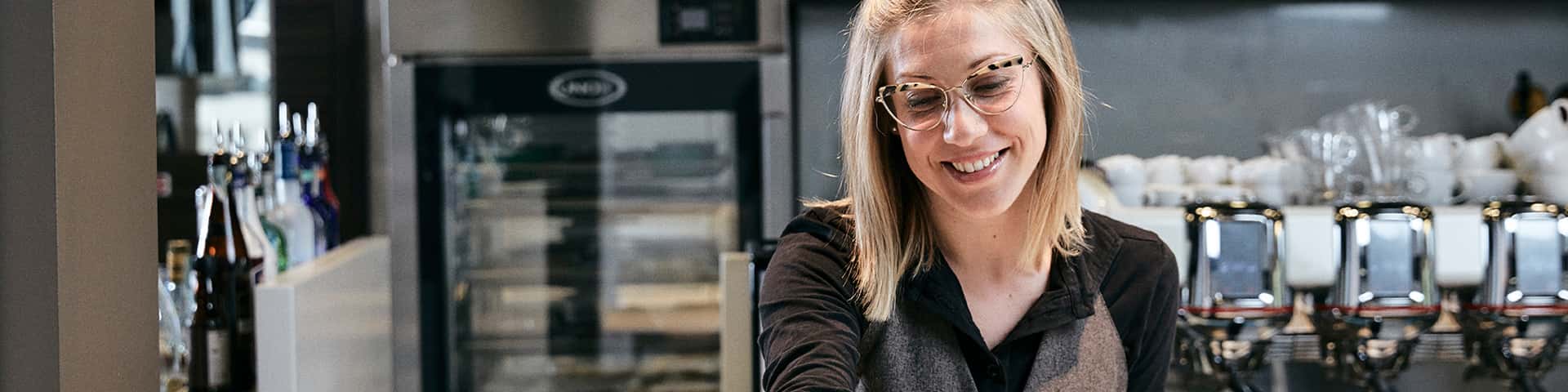 Smiling woman with glasses adding herbs to a dish of risotto, coffee machines, dishes of food and Unox Evereo can be seen behind her.