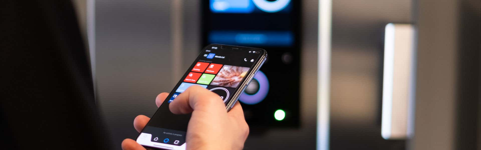Man in black chef jacket holding a smart-phone pressing some buttons, facing towards an unox speed-x speed oven, controlling the oven with his phone.