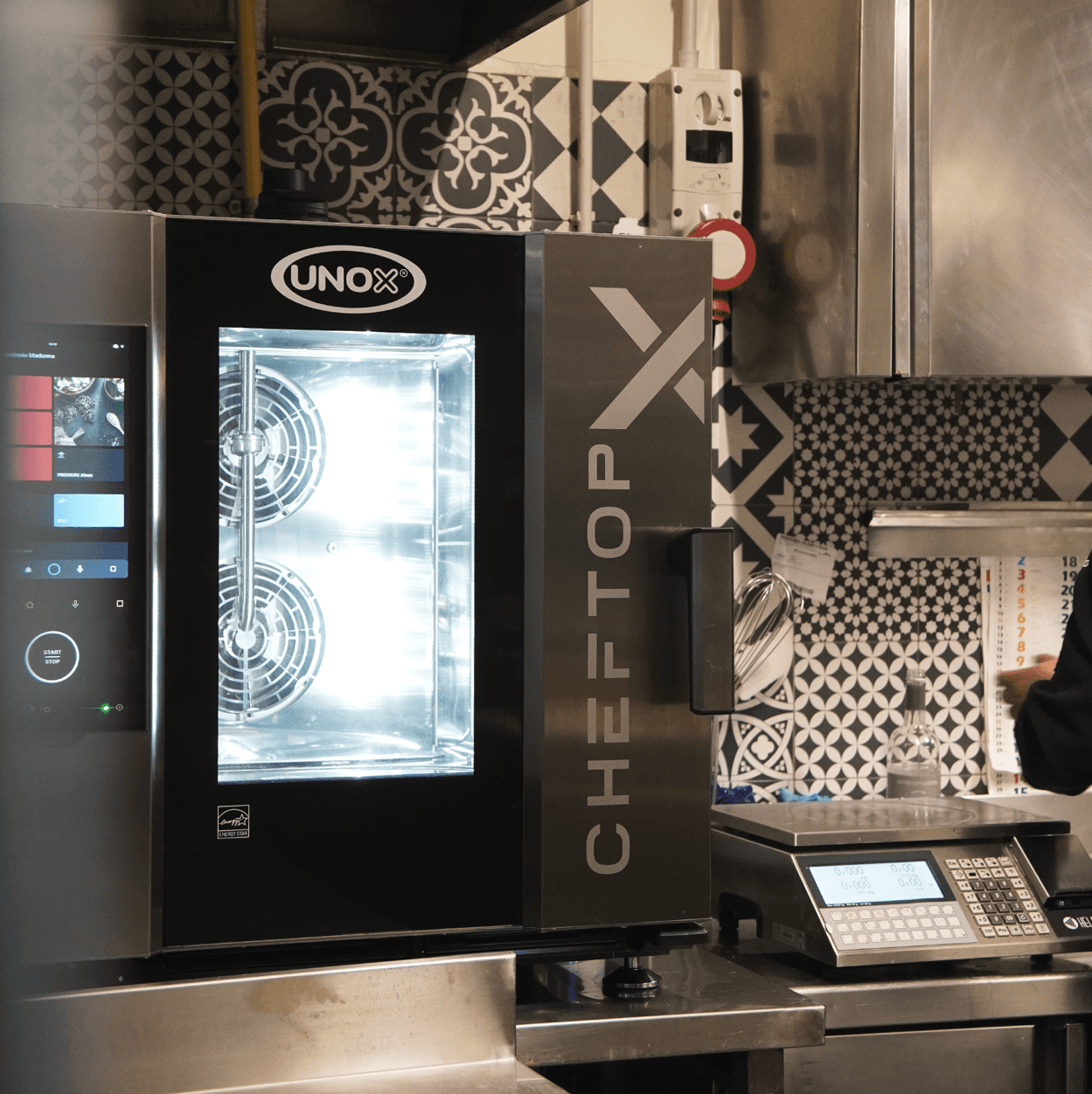 Intelligenter Unox Kombidämpfer mit eingeschaltetem Touchscreen-Display, platziert auf einer Theke in einer Profiküche neben einer Waage