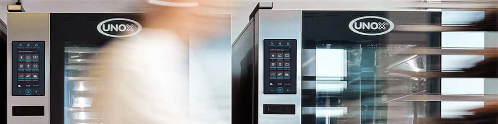 Cocina con dos hornos de carro unox; un cocinero con gorro blanco pasa empujando un carrito, creando desenfoque de movimiento.