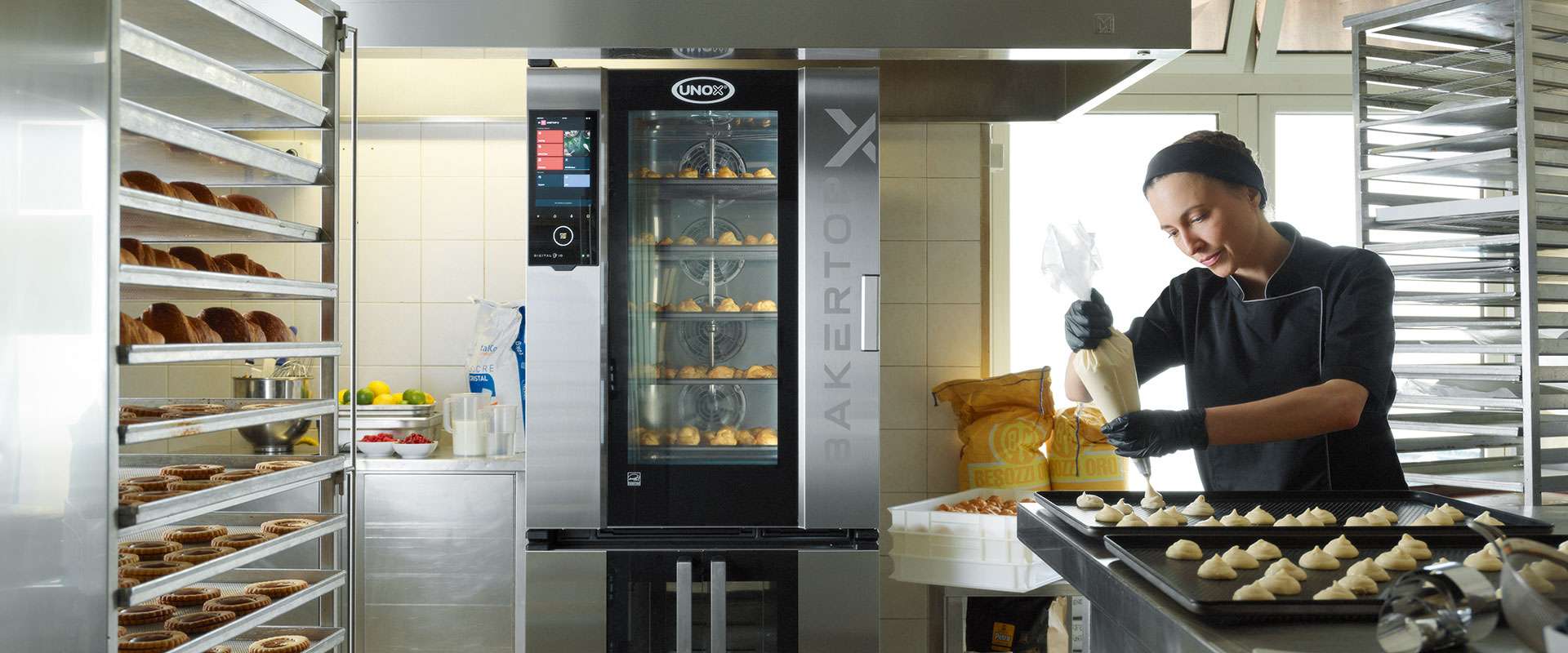 A baker in a kitchen, standing near an Unox combi oven with pastries inside, holds a piping bag. Pastries are displayed on a nearby trolley.