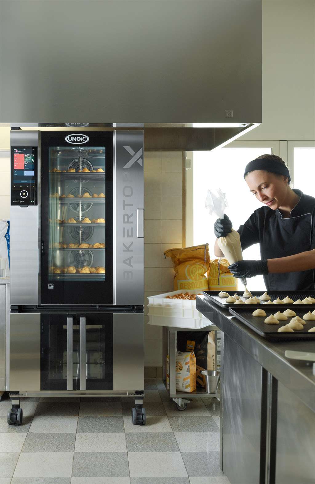 A baker in a kitchen, standing near an Unox combi oven with pastries inside, holds a piping bag. Pastries are displayed on a nearby trolley.