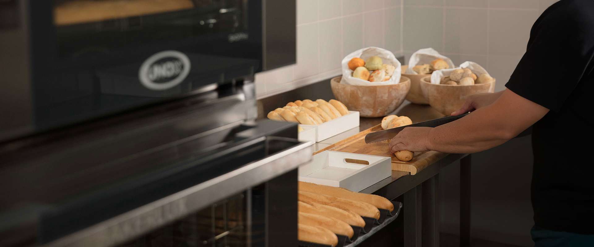 Bakery worker prepares baguettes and pastries beside a Unox BAKERLUX SHOP.Pro commercial oven, with trays of bread and bowls of dough creating a warm, busy kitchen scene.