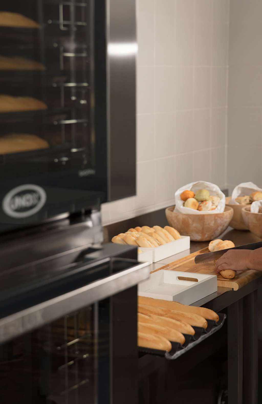 Bakery worker prepares baguettes and pastries beside a Unox BAKERLUX SHOP.Pro commercial oven, with trays of bread and bowls of dough creating a warm, busy kitchen scene.