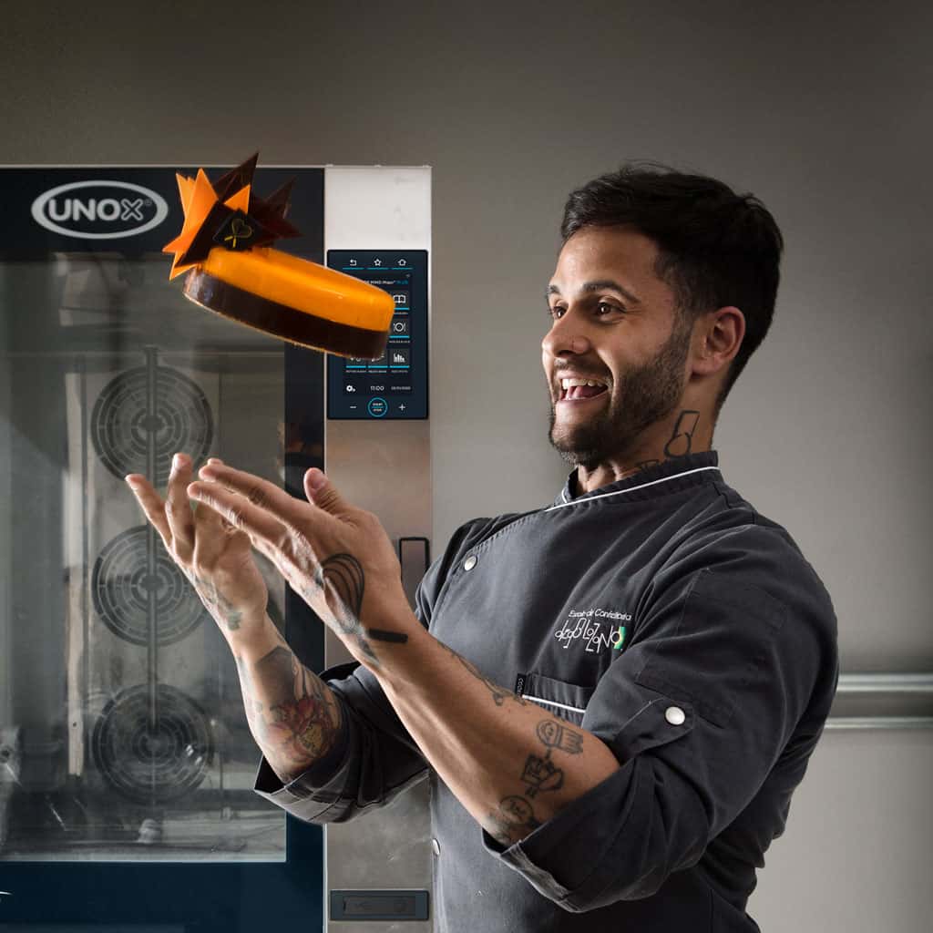Smiling male chef throwing an orange cake into the air in front of an unox baking oven.