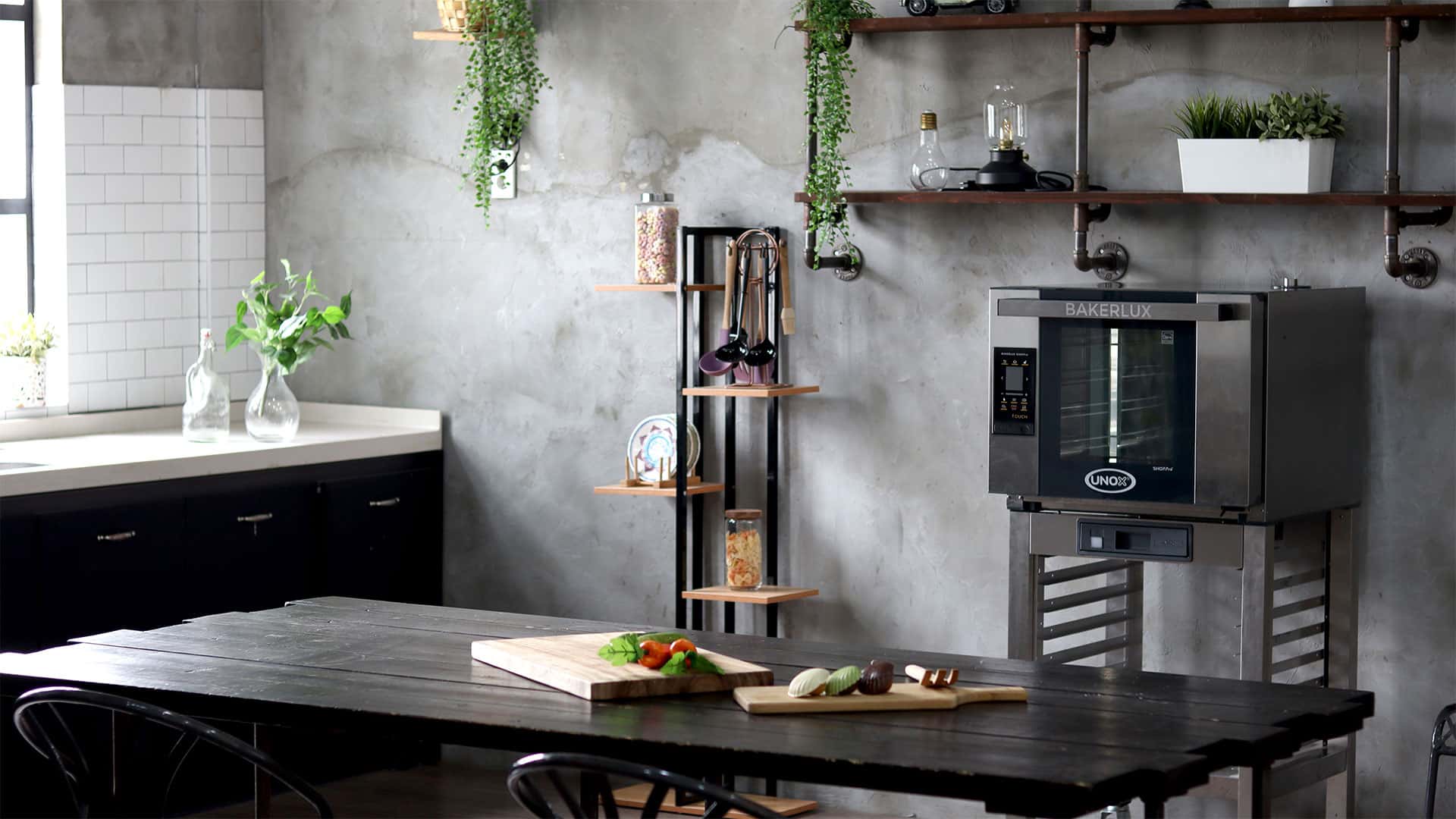 Grey home kitchen with green plants and large black table in centre. Wooden boards with food on the table. Next to the table is an Unox baking oven.