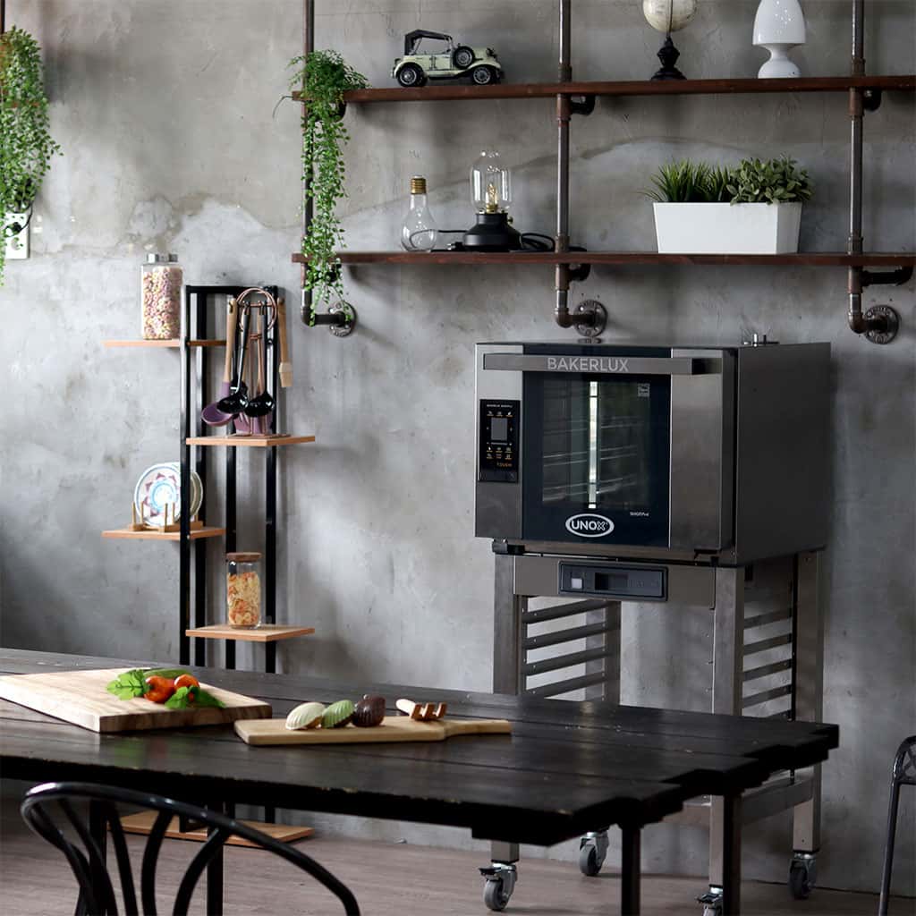 Grey home kitchen with green plants and large black table in centre. Wooden boards with food on the table. Next to the table is an Unox baking oven.