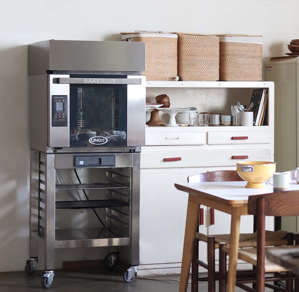 Home kitchen with dining table and chairs, white cupboards filled with cake stands and mugs, next to an unox baking oven on a stand with wheels.