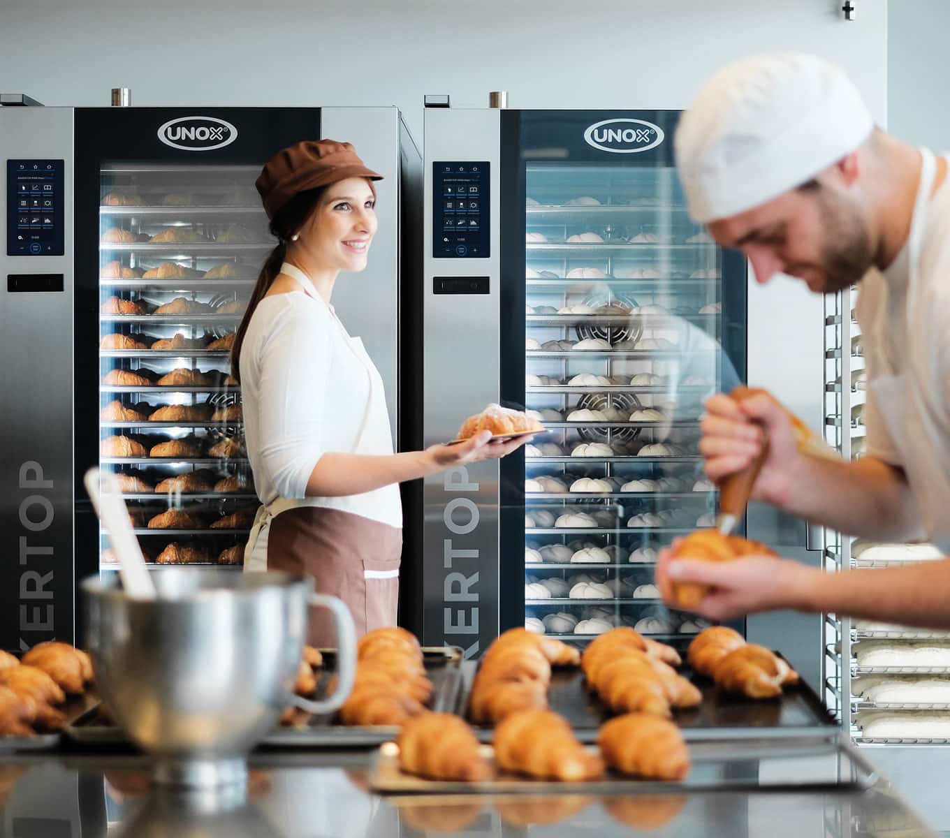 To operatører forbereder croissanter i laboratoriet med to Unox kombiovne med vogn, fuldt lastet, i baggrunden