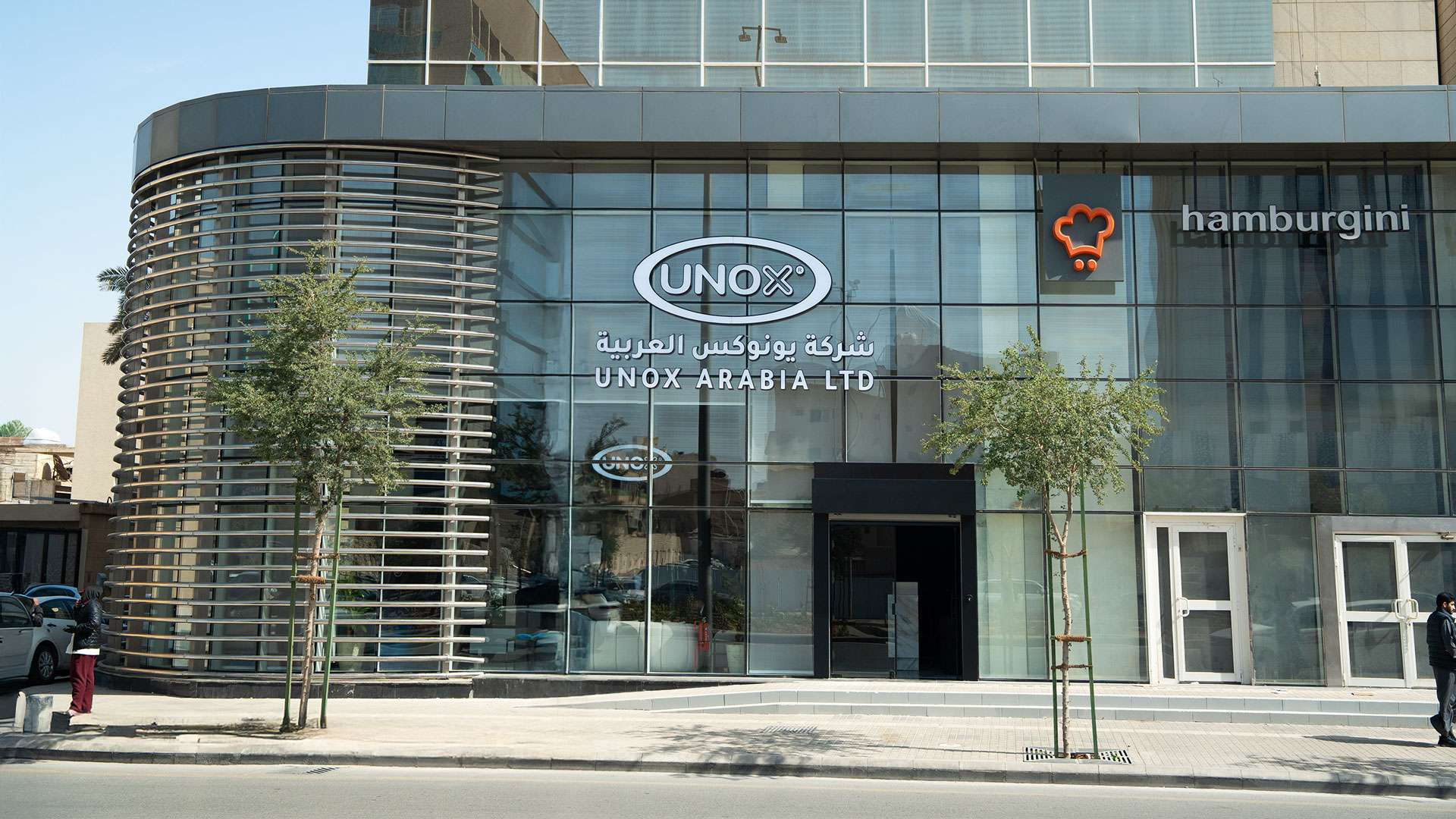 Glass-fronted building with "UNOX Arabia Ltd" signage in English and Arabic. Nearby, a logo and "hamburgini" text are visible. Trees line the sidewalk.