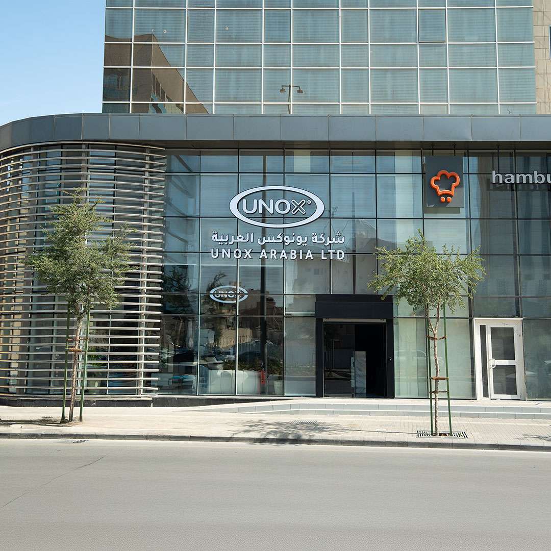 Glass-fronted building with "UNOX Arabia Ltd" signage in English and Arabic. Nearby, a logo and "hamburgini" text are visible. Trees line the sidewalk.