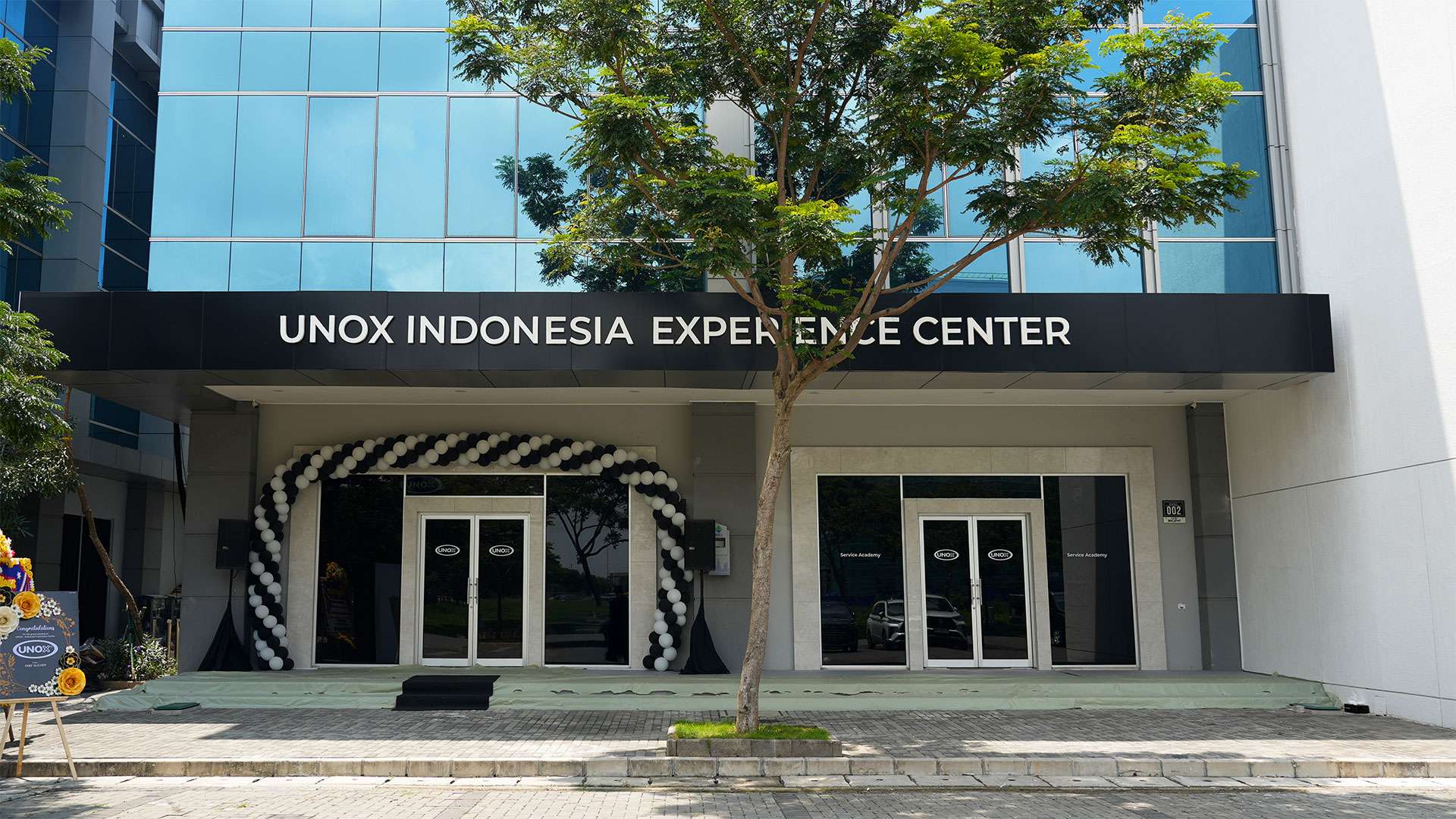 Facade of the UNOX Indonesia Experience Center featuring glass doors, a black sign with white lettering, and a decorative arch of black and white balloons.