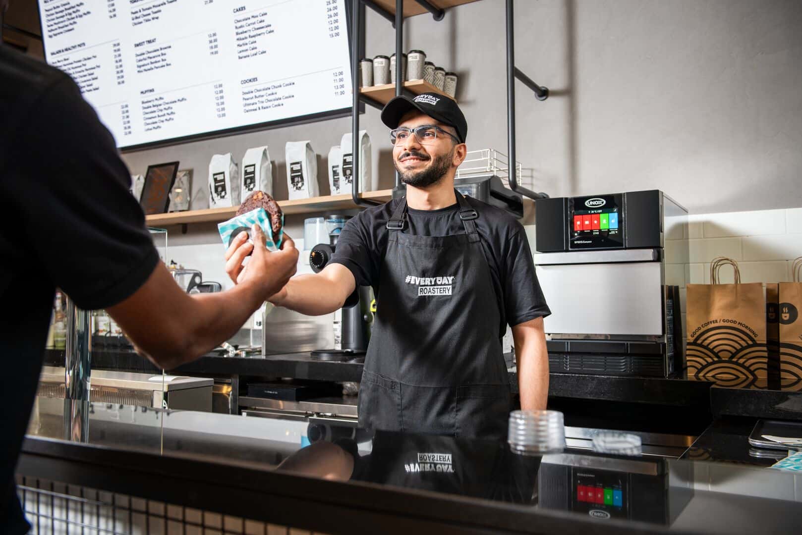 Barista serves a hot cookie to a customer, with a Unox speed compact oven visible on the countertop.